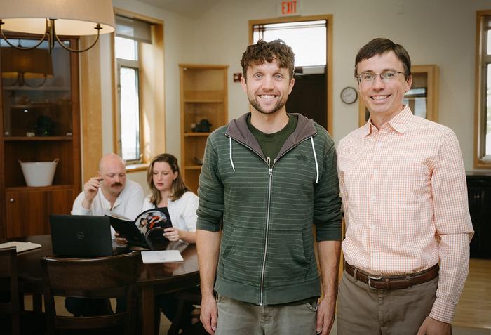 Two men are standing in a living room setting, smiling and looking into the camera. In the background, a man and a woman are sitting at a table reading a brochure.