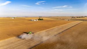 A combine harvester moves through a large, golden field under a blue sky; farm buildings are visible in the background