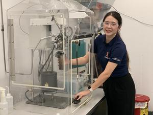 A woman in a blue t-shirt and lab glasses is standing in a laboratory demonstrating a process