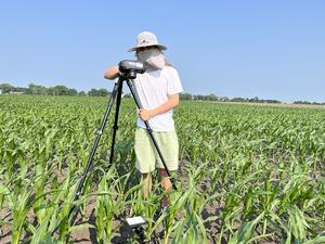 A man standing with a camera on a tripod in a maize field