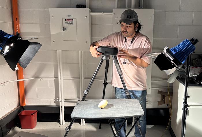 A man in a lab setting holding a camera over a sweet potato on a table