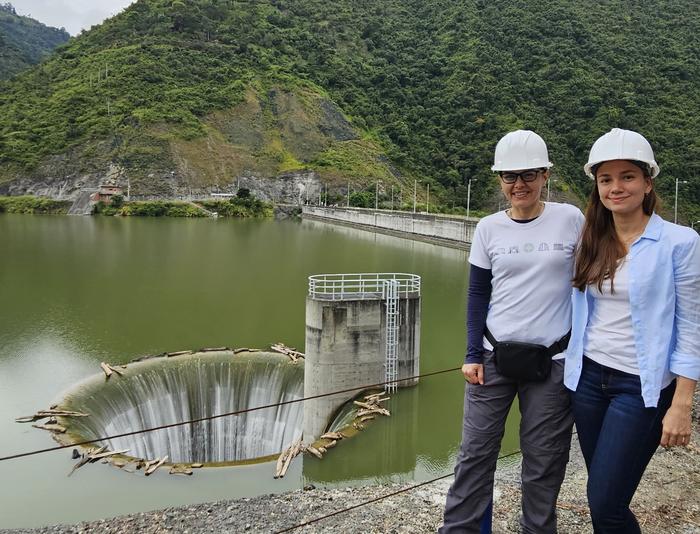 Two women wearing hard hats stand near a reservoir with a visible morning glory spillway and surrounding green hills in the background.